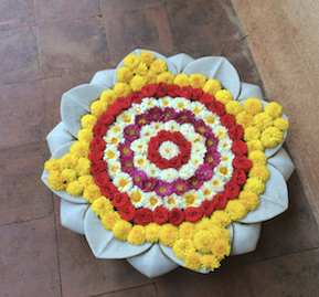 A mandala of marigold, rose and white petals arranged on a pale dish on a dark wooden floor
