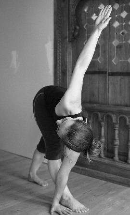 Black and white: a standing forward fold with one leg lifted high, in front of a carved wooden panel